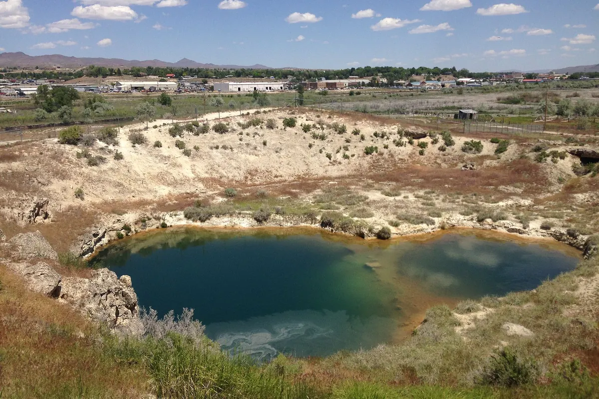 Blue-green Elko Hot Hole pool ringed by pale mineral crust, Elko buildings in the distance